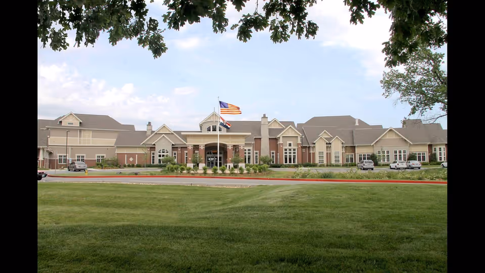 Front exterior view of Aberdeen Heights facility with a large lawn in the foreground, an American flag and another flag flying on flagpoles, and several cars parked near the building under a partly cloudy sky.