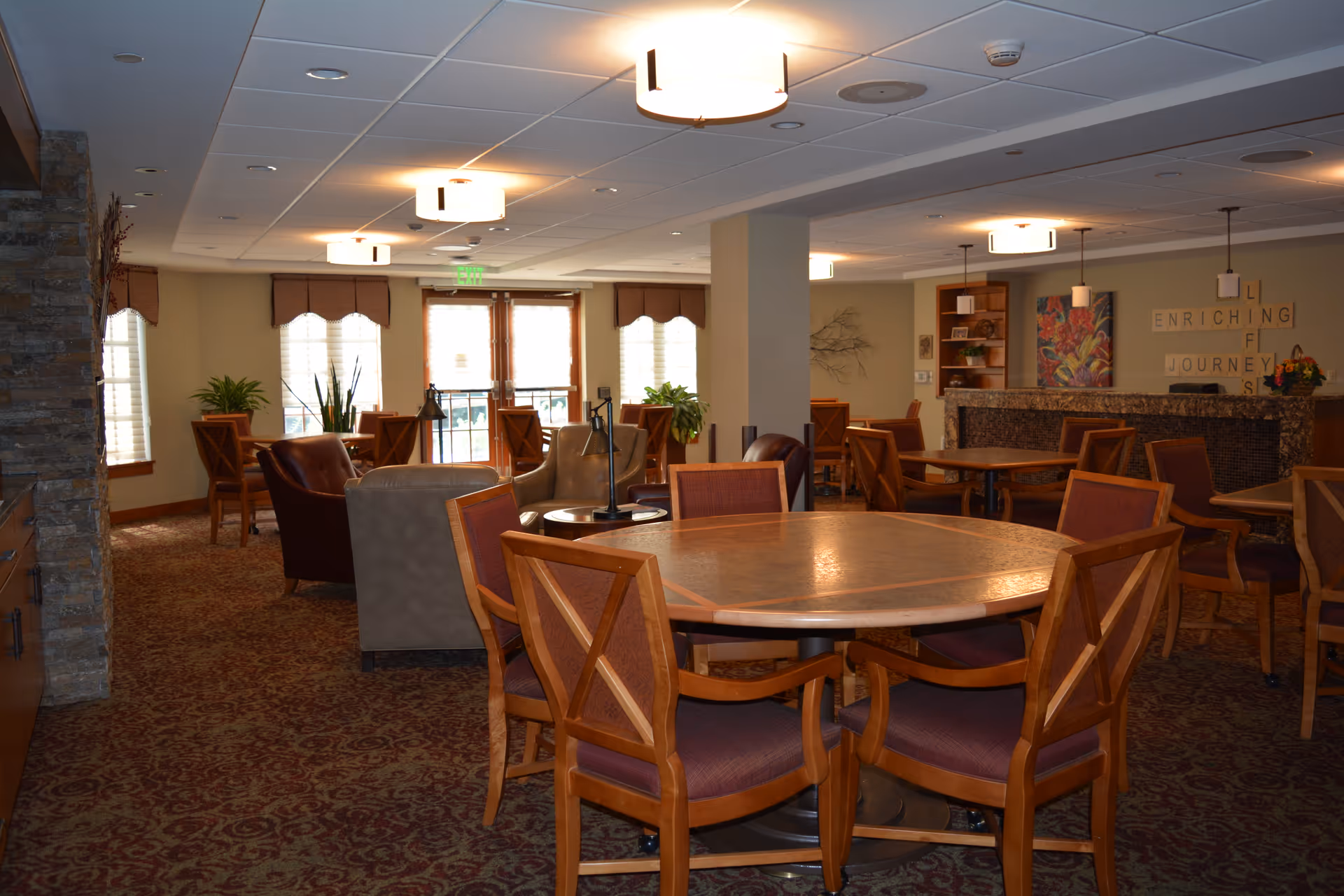 A spacious and well-lit common area with multiple round wooden tables and chairs arranged for seating. The room features carpeted flooring with a patterned design, several armchairs, large windows with blinds, and decorative plants. The walls have artwork and a sign that reads 'ENRICHING JOURNEYS'. Ceiling lights provide warm illumination.
