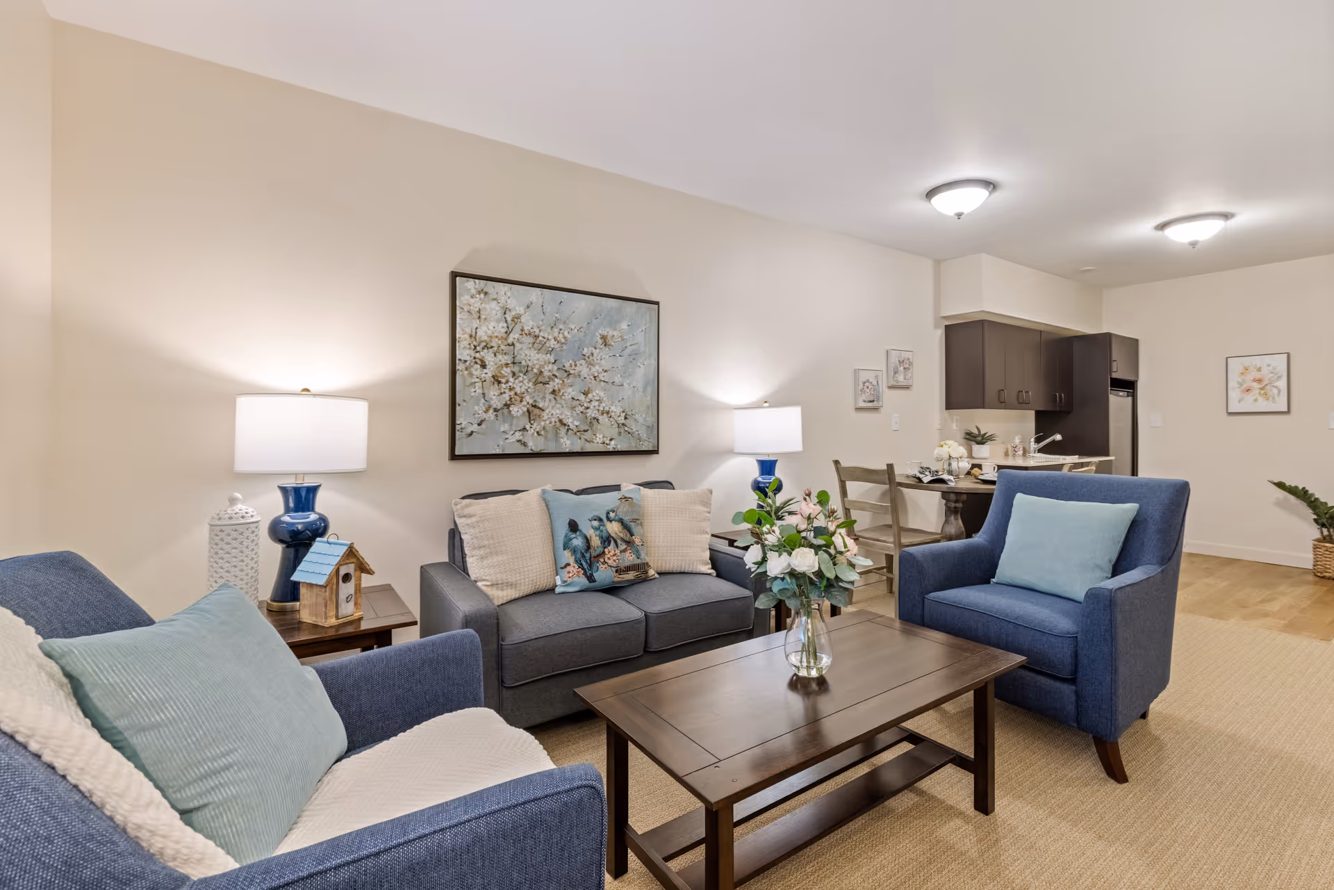 A cozy living room with two blue armchairs and a blue sofa adorned with cushions. A wooden coffee table with a vase of flowers sits in the center. Two table lamps with white shades are on side tables beside the sofa. The background shows a small dining area with a wooden table and chairs, and a kitchen with dark cabinets and a refrigerator. The walls are decorated with framed artwork.