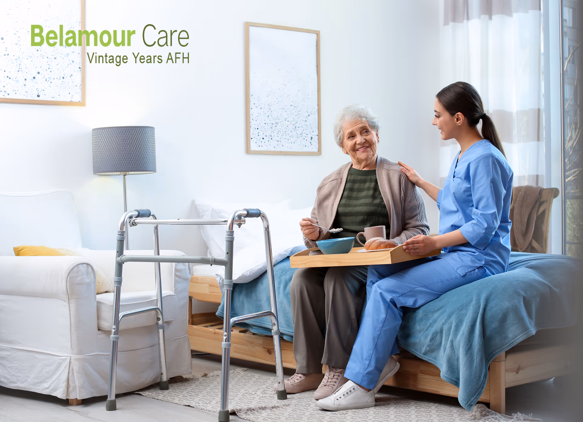 An elderly woman sitting on a bed with a tray of breakfast food, accompanied by a caregiver in blue scrubs who is sitting beside her and gently touching her shoulder. A walker is positioned in front of the bed. The room is bright and cozy with a white armchair, a floor lamp, framed artwork on the wall, and sheer curtains letting in natural light. The text 'Belamour Care Vintage Years AFH' is visible on the wall.