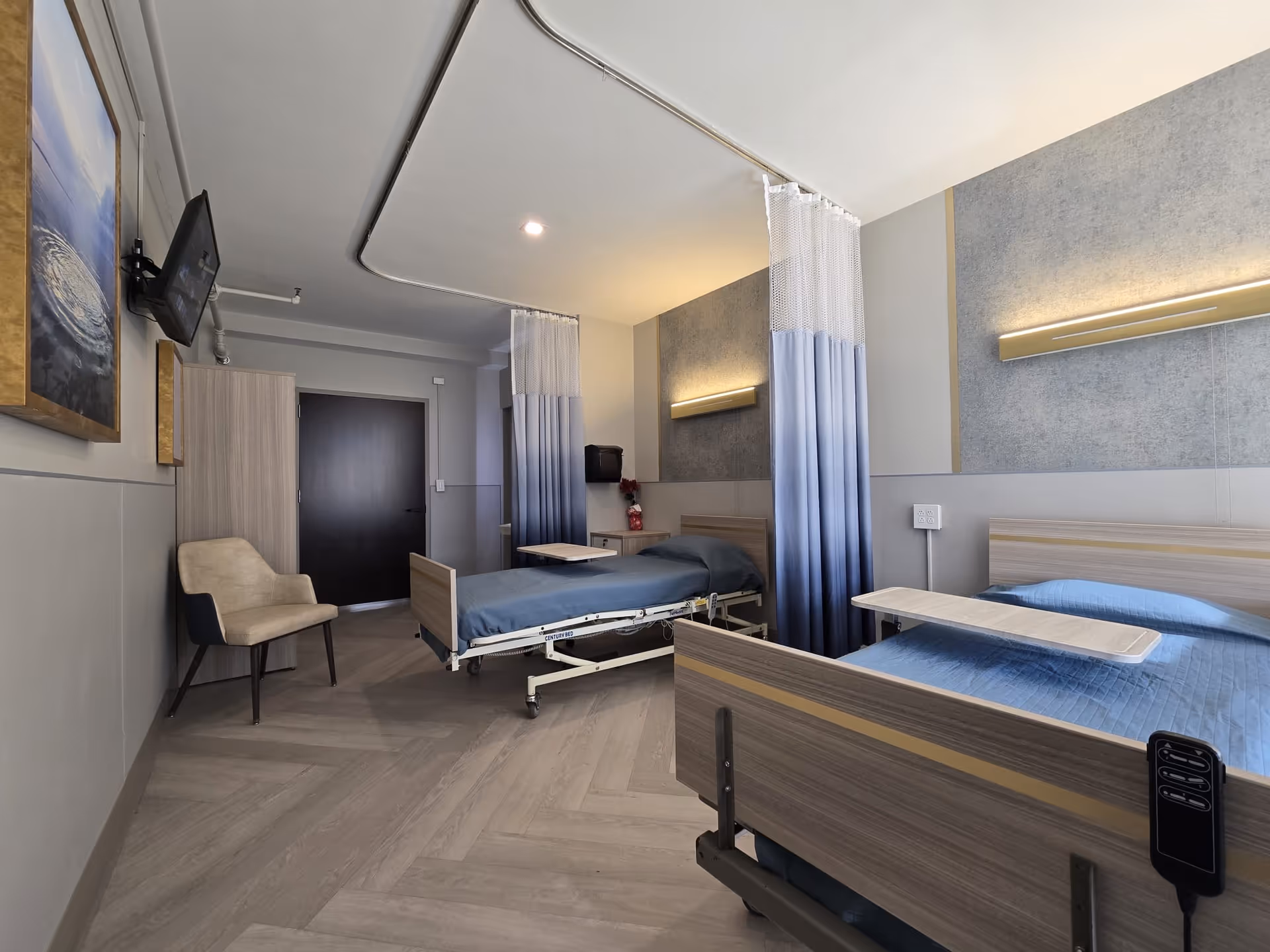 A clean and modern hospital-style room with two adjustable beds separated by a blue and white privacy curtain. Each bed has a light blue bedspread and an overbed table. The room features a beige armchair, a wall-mounted TV, a wooden wardrobe, and soft lighting fixtures above the beds. The floor has a light wood pattern, and the walls are painted in neutral tones with a textured accent behind the beds.