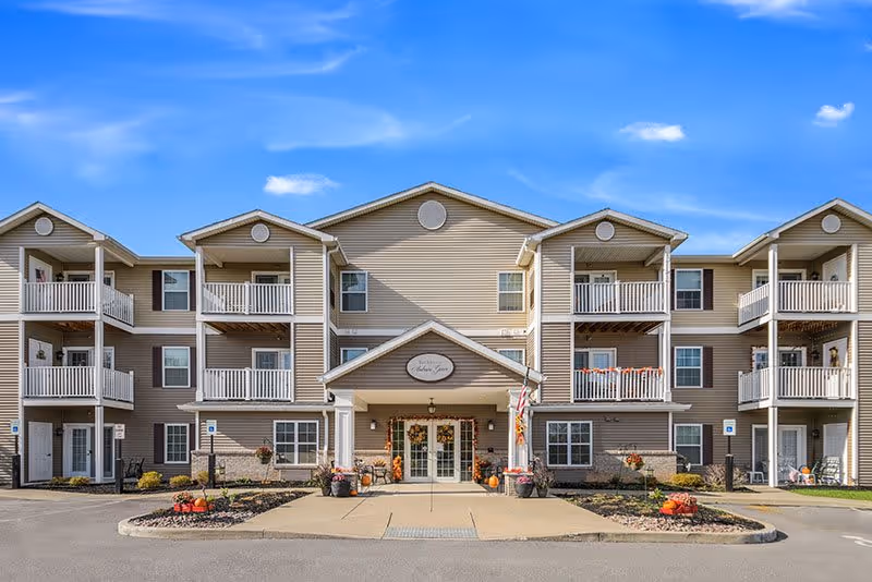Front exterior view of a three-story senior living facility building with beige siding and white balconies under a clear blue sky. The entrance is decorated with autumn-themed decorations including pumpkins and flowers.