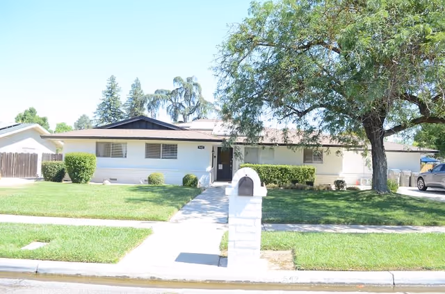 Single-story residential building with a light-colored exterior, a front lawn with green grass, bushes, and a large tree on the right side. A concrete walkway leads from the sidewalk to the front door. A white mailbox is positioned near the sidewalk. A car is parked on the driveway to the right of the house.