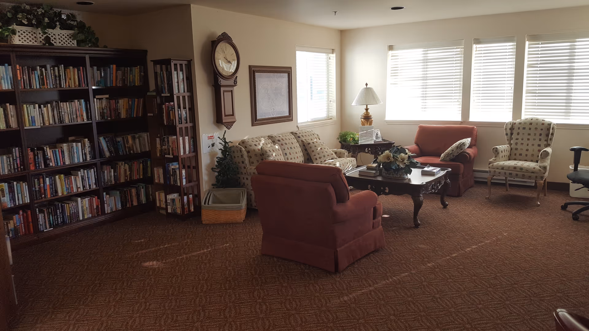 A cozy senior living room with a patterned sofa, two red armchairs, a patterned armchair, and a wooden coffee table with a flower arrangement. There is a large bookshelf filled with books on the left side, a wall clock, framed artwork, and windows with blinds letting in natural light. The room has a carpeted floor and a table lamp on a side table.