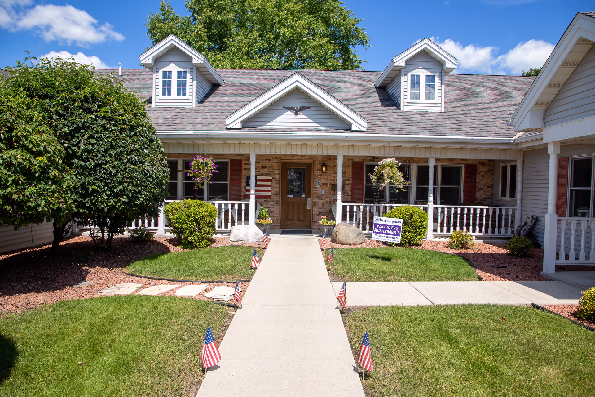 Front exterior view of a single-story assisted living facility building with a gray shingled roof, white trim, and a covered porch. The entrance is centered with a wooden door, flanked by windows with red shutters. There are hanging flower baskets on the porch, small American flags lining the walkway, and a sign on the lawn promoting an Alzheimer's walk event. The sky is blue with some clouds, and there are trees and shrubs around the building.