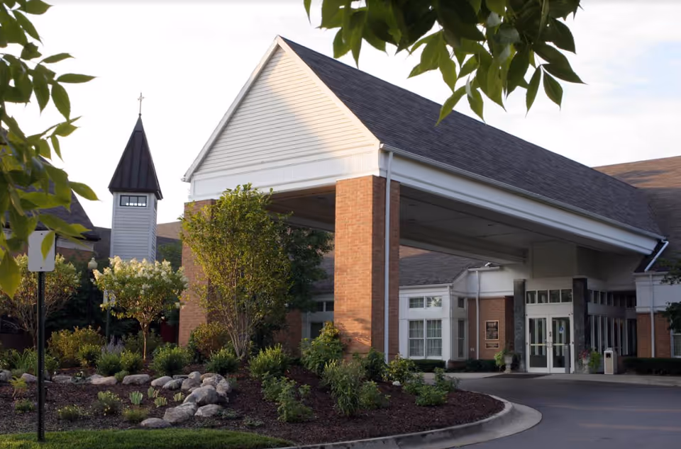 Entrance of The Orchards at Canterbury-on-the-Lake facility showing a covered driveway supported by brick pillars, surrounded by landscaped greenery and trees, with a building and a small tower with a cross in the background.