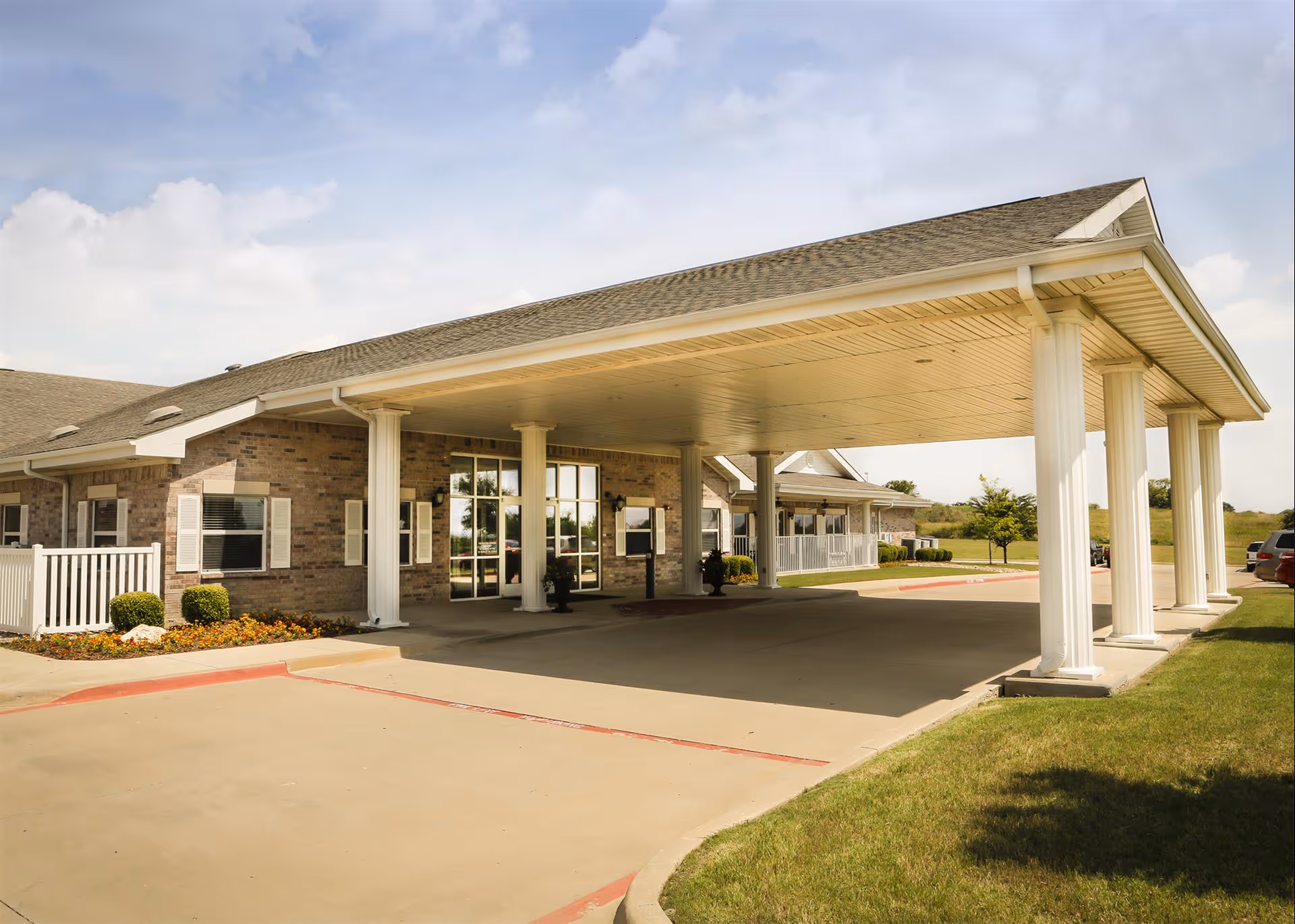 Exterior view of Bethesda Gardens Fort Worth showing the covered entrance with white columns, brick walls, windows with shutters, and a driveway with parking spaces. The sky is partly cloudy and there is green grass and some landscaping around the building.