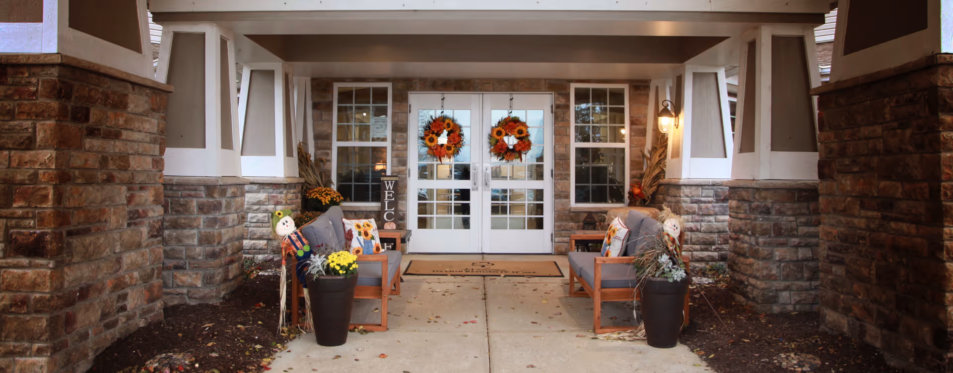 Entrance to a building with double glass doors decorated with autumn-themed wreaths. Two cushioned benches with fall-themed pillows and potted plants with scarecrows flank the walkway. Stone and white pillars frame the entrance, and a welcome mat is placed in front of the doors.