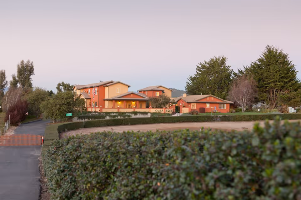 Exterior view of Casa Rosa Elder Care facility during dusk, showing multiple connected buildings with warm lighting, surrounded by trees and bushes, with a driveway and a hedge in the foreground.