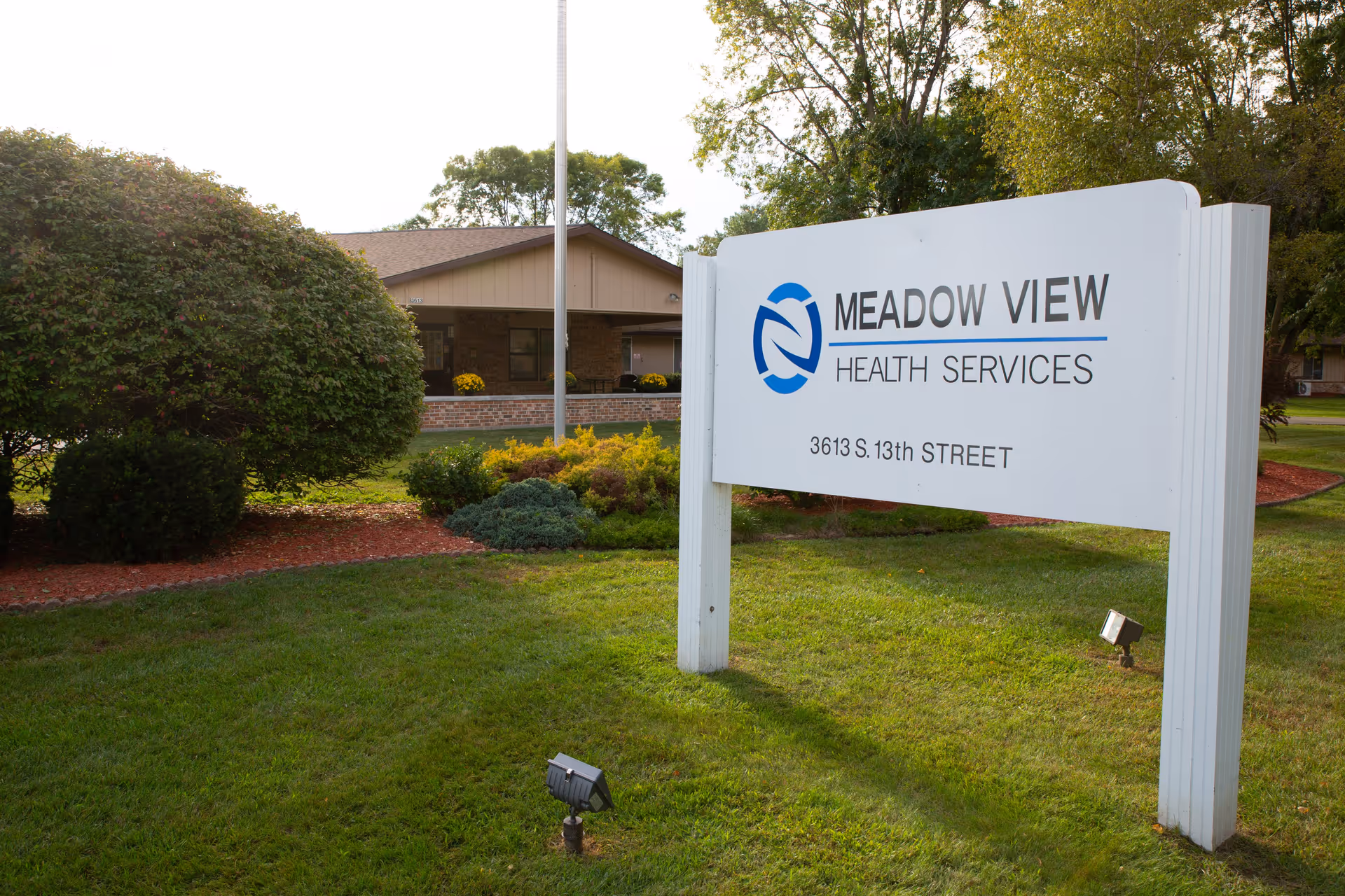 Outdoor view of Meadow View Health Services sign on a grassy lawn with bushes and a building in the background.