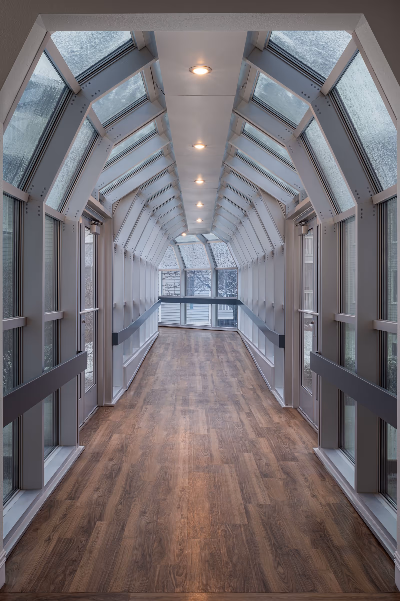 A long, enclosed hallway with large glass windows and a glass ceiling on both sides, featuring wooden flooring and ceiling lights evenly spaced along the center.