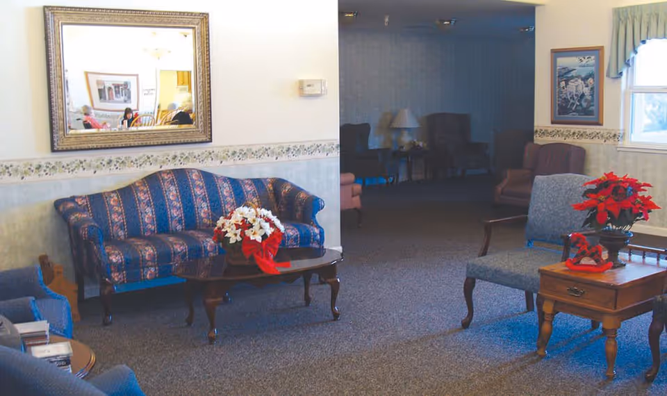 A cozy living room area with a patterned blue sofa, a wooden coffee table with a floral arrangement, and several upholstered chairs. There is a large mirror on the wall reflecting part of the room, a window with curtains, and framed artwork on the walls. The room has carpeted flooring and soft lighting.