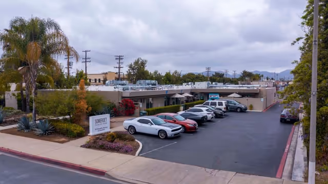 Parking lot in front of a single-story building with several parked cars and landscaping including palm trees and bushes. A sign near the sidewalk reads 'Somerset Subacute Rehab'. The sky is cloudy and there are power lines and trees in the background.
