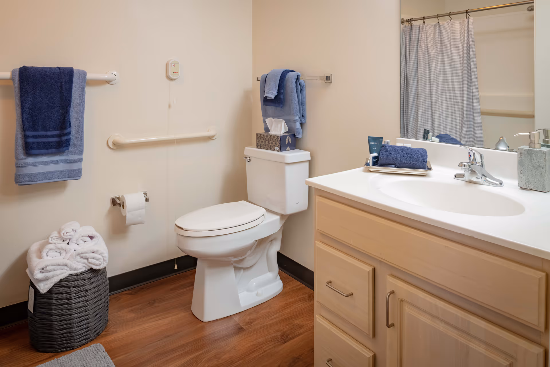 A clean and accessible bathroom featuring a white toilet with a tissue box and folded blue towels on top, a wooden vanity with a white sink and silver faucet, a large mirror above the sink, a shower with a curtain in the background, blue towels hanging on wall-mounted racks, a basket filled with rolled white towels on the wooden floor, and grab bars on the walls for support.