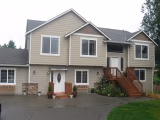 Exterior front view of a two-story beige house with white trim, multiple windows, a wooden staircase leading to the main entrance, and a driveway in front. There are some plants and greenery around the entrance area.