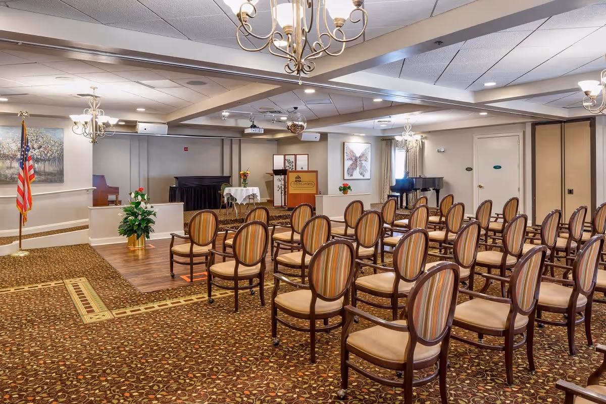 A well-lit meeting or event room with rows of wooden chairs featuring striped upholstery arranged facing a small stage area. The stage has a podium with a sign that reads 'Aston Gardens At Parkland Commons,' a piano, and a small table with flowers. The room has patterned carpet, chandeliers, an American flag, and framed artwork on the walls.