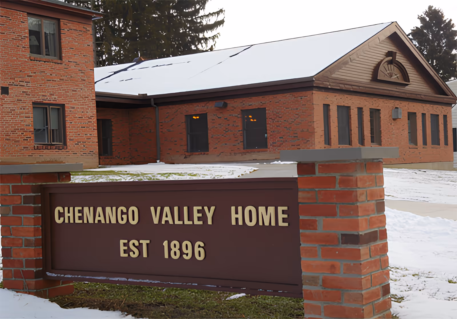 Exterior view of a brick building with a snow-covered roof and a sign in front that reads 'Chenango Valley Home EST 1896'. The ground is partially covered with snow and there are trees in the background.