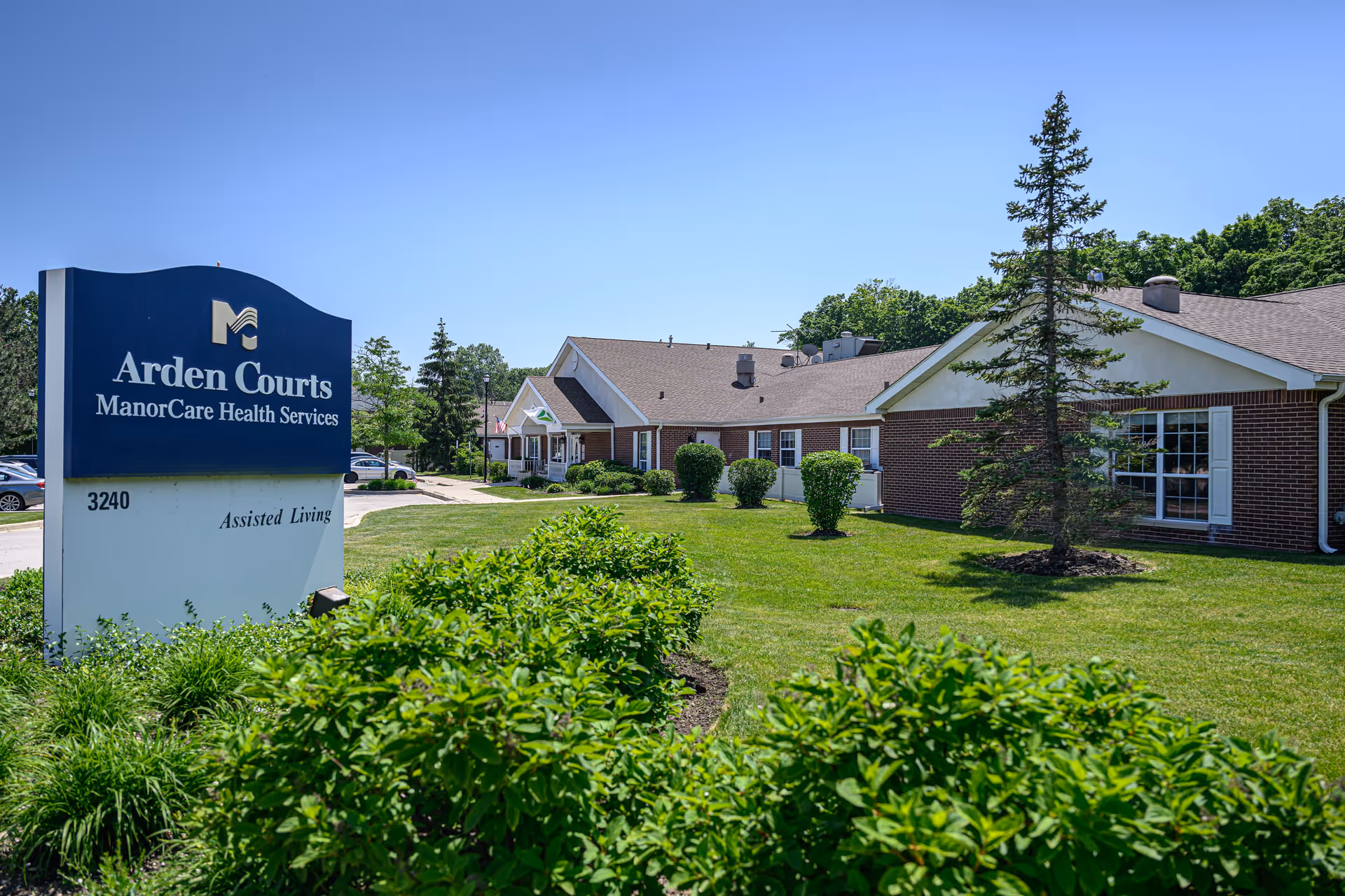 Exterior view of Arden Courts ManorCare Health Services assisted living facility with a large sign in the foreground and a brick building surrounded by green grass, bushes, and trees under a clear blue sky.