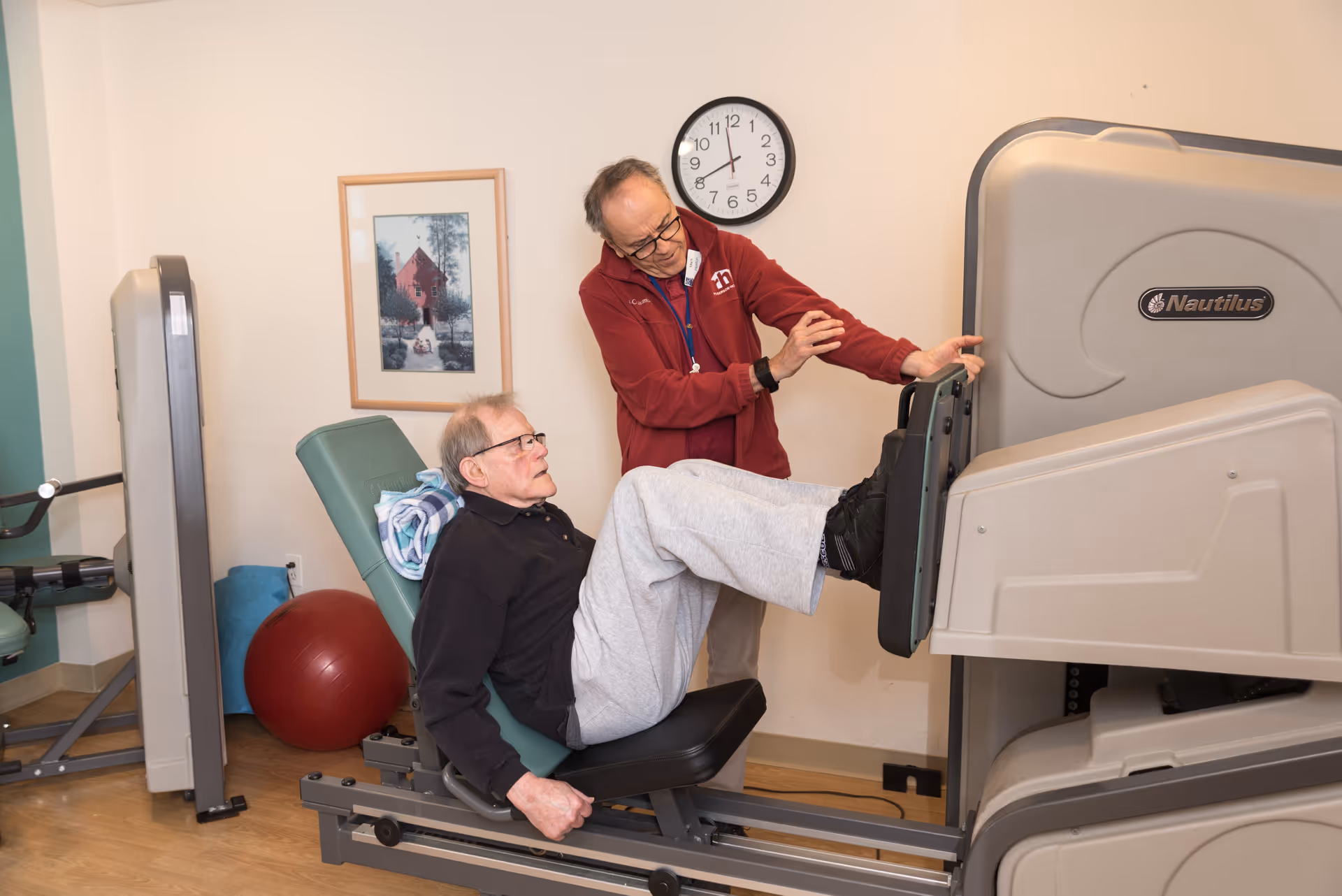 An elderly man is seated on a leg press exercise machine in a fitness room, pushing against the footplate. A caregiver or trainer stands beside him, providing assistance and encouragement. The room has light-colored walls, a clock, a framed picture, and exercise equipment including a red exercise ball.