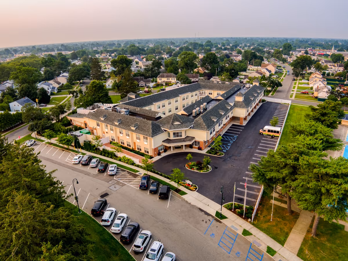 Aerial view of the Village Green senior living facility showing a large, multi-story building with a U-shaped layout surrounded by parking lots and greenery. The surrounding neighborhood with houses and trees extends into the distance under a hazy sky.