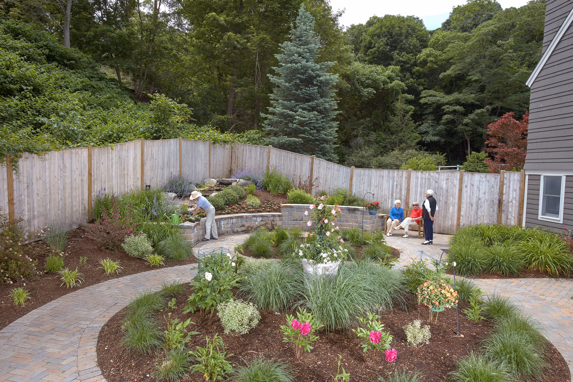 A landscaped garden area with a circular flower bed in the center, surrounded by a paved walkway. One person wearing a hat is tending to plants near a wooden fence, while three elderly individuals are sitting and standing nearby, enjoying the outdoor space. The area is bordered by trees and greenery, with part of a building visible on the right.