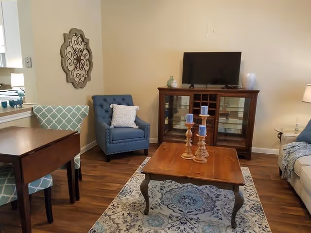 A cozy living room with a wooden coffee table holding three blue candles on wooden holders, a patterned area rug, a blue armchair with a white pillow, a wooden cabinet with a flat-screen TV on top, and a beige sofa partially visible on the right. There is a decorative wall piece above the armchair and a small table with a lamp on the right side.