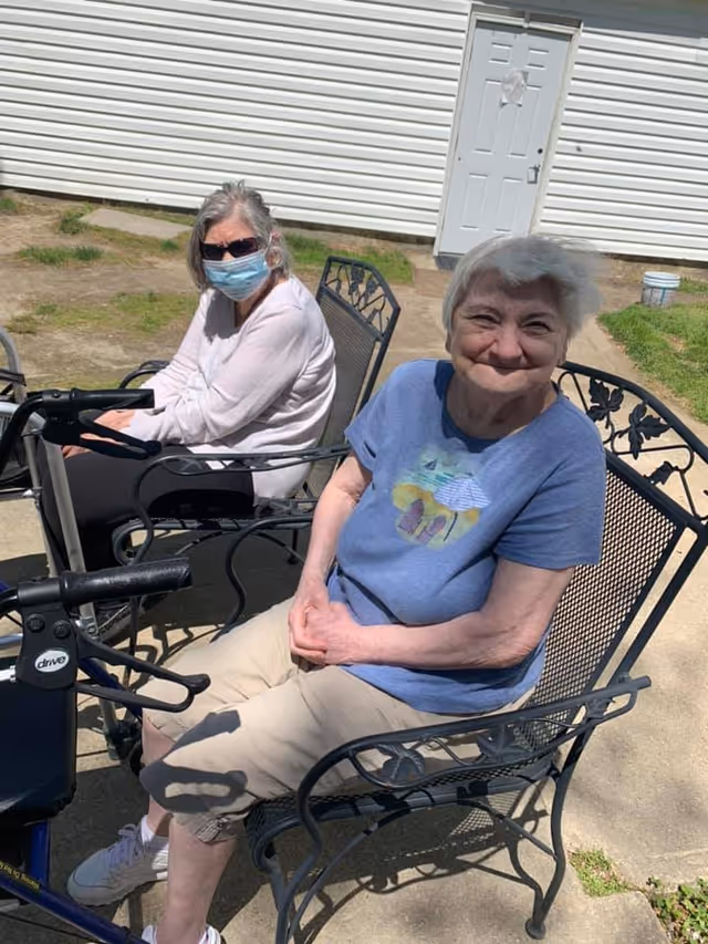 Two elderly women sitting outside on metal chairs near a white building. One woman is wearing a blue shirt and beige shorts, smiling at the camera. The other woman is wearing a light pink long-sleeve shirt, black pants, sunglasses, and a face mask. There are mobility aids visible near them.