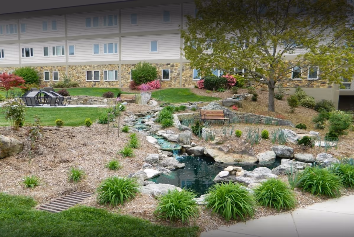 Outdoor garden area at Carolina Village featuring a small pond with rocks and a water feature, surrounded by green plants and mulch. There are benches and a seating area with chairs on a stone patio. The background shows a multi-story building with stone and siding exterior and several windows.