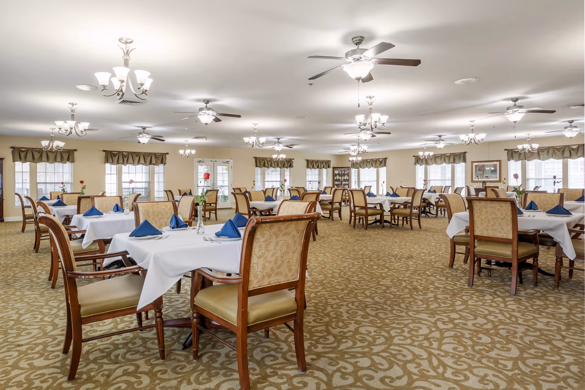 A spacious dining room with multiple tables covered in white tablecloths, each set with blue folded napkins and a single red rose in a vase. The room features patterned carpet, beige walls, multiple windows with valances, ceiling fans, and chandeliers providing bright lighting.