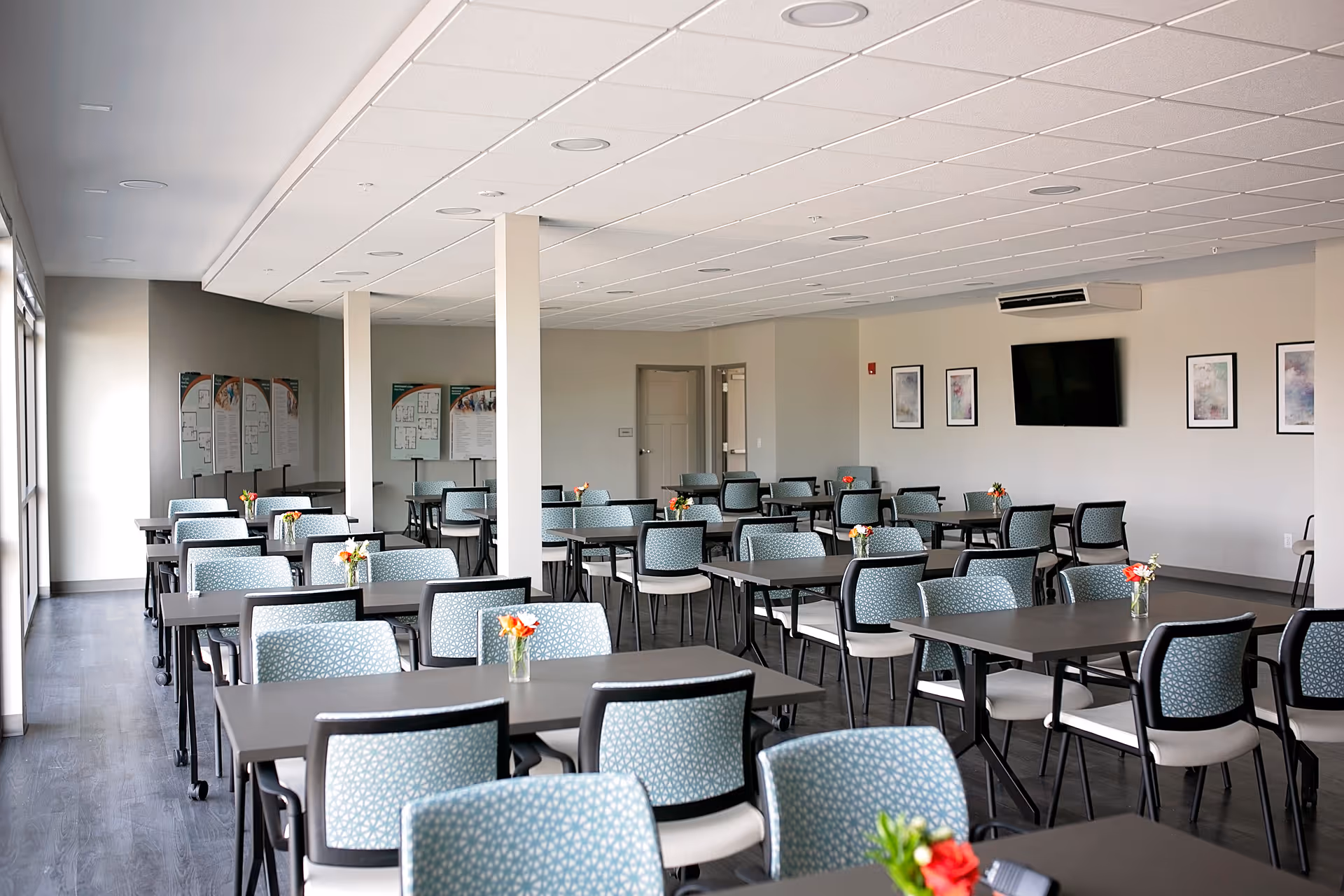 A spacious dining room with multiple tables and chairs arranged neatly. Each table has a small vase with flowers. The room has large windows on one side allowing natural light to enter, a flat-screen TV mounted on the wall, and framed artwork. The flooring is dark wood, and the ceiling has recessed lighting.