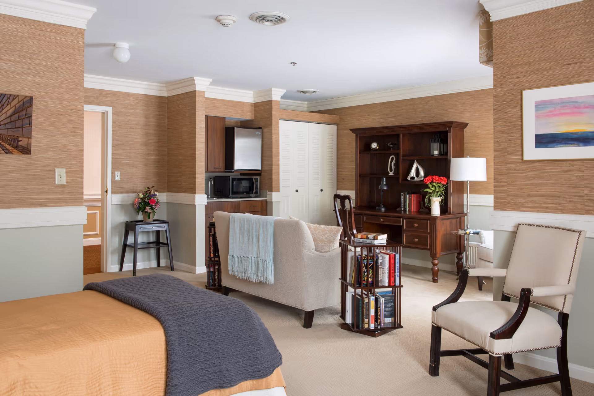 A cozy senior living facility room featuring a bed with a mustard-colored quilt and gray blanket, a beige upholstered armchair, a small sofa with a light blue throw, a wooden bookshelf with decorative items and books, a small rotating bookshelf filled with books, a kitchenette area with a microwave and mini fridge, and a side table with a vase of flowers. The walls have a textured brown wallpaper with white trim, and there is a colorful framed painting on the wall.