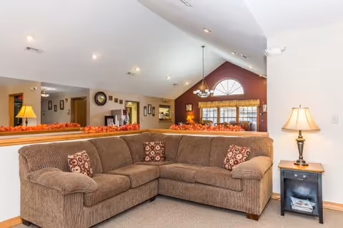 A cozy living room area featuring a large brown sectional sofa with patterned throw pillows. The room has a high vaulted ceiling with recessed lighting and a hanging chandelier. A large window with a half-circle top lets in natural light, and the walls are painted in warm tones including a deep red accent wall. There is a small side table with a lamp next to the sofa, and decorative autumn-themed garland is placed on the half wall behind the sofa.