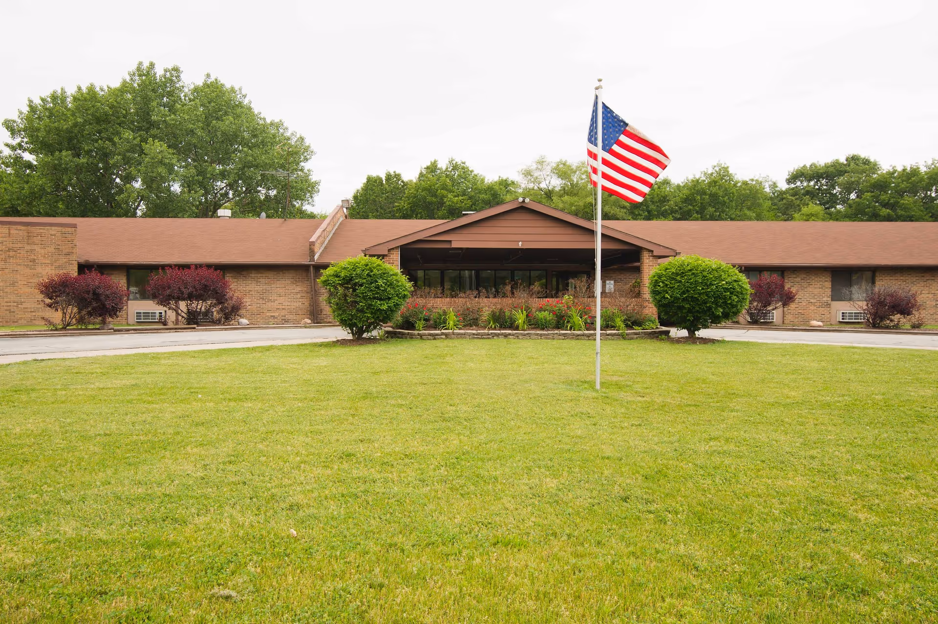 Front exterior view of a single-story brick building with a brown roof, surrounded by green grass and bushes. An American flag on a flagpole is prominently displayed in the center of the lawn. Trees are visible in the background under an overcast sky.
