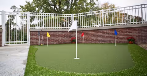 Outdoor putting green with four small flags in yellow, white, red, and blue, surrounded by a white metal fence and a brick wall with potted plants, with trees visible in the background.