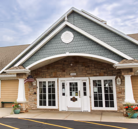 Front entrance of a senior living facility with a peaked roof, stone and siding exterior, large white-framed windows, a white door, and two potted plants on either side of the entrance. An American flag is mounted on the left pillar.
