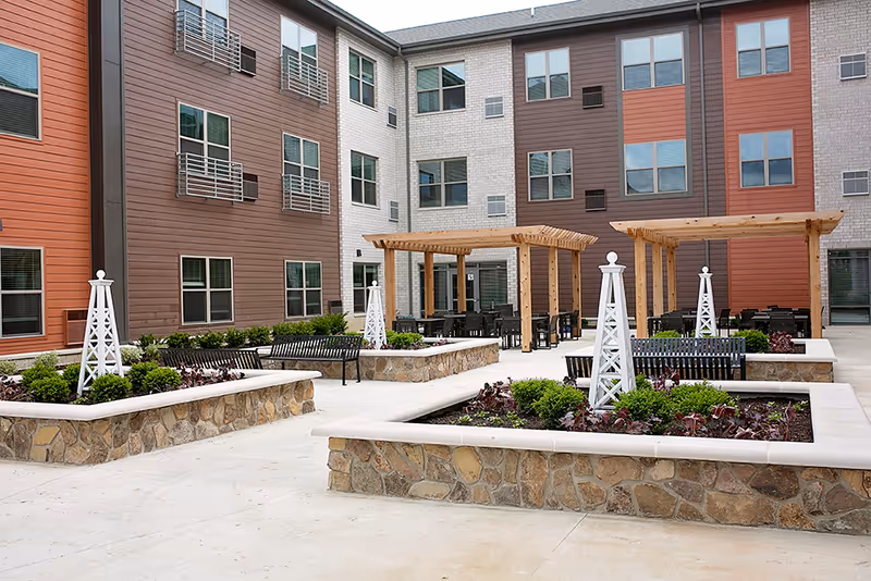 Outdoor courtyard area of a senior living facility with raised stone flower beds containing plants and white decorative trellises. There are black benches around the flower beds and wooden pergolas with tables and chairs underneath. The building surrounding the courtyard has multiple windows and is clad in brown, white, and orange siding.