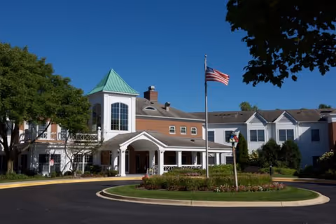Exterior view of a senior living facility building with a circular driveway and landscaped roundabout featuring flowers and greenery. The building has a white facade with brick accents, a green peaked roof tower, and an American flag flying on a flagpole in front. The sky is clear and blue.