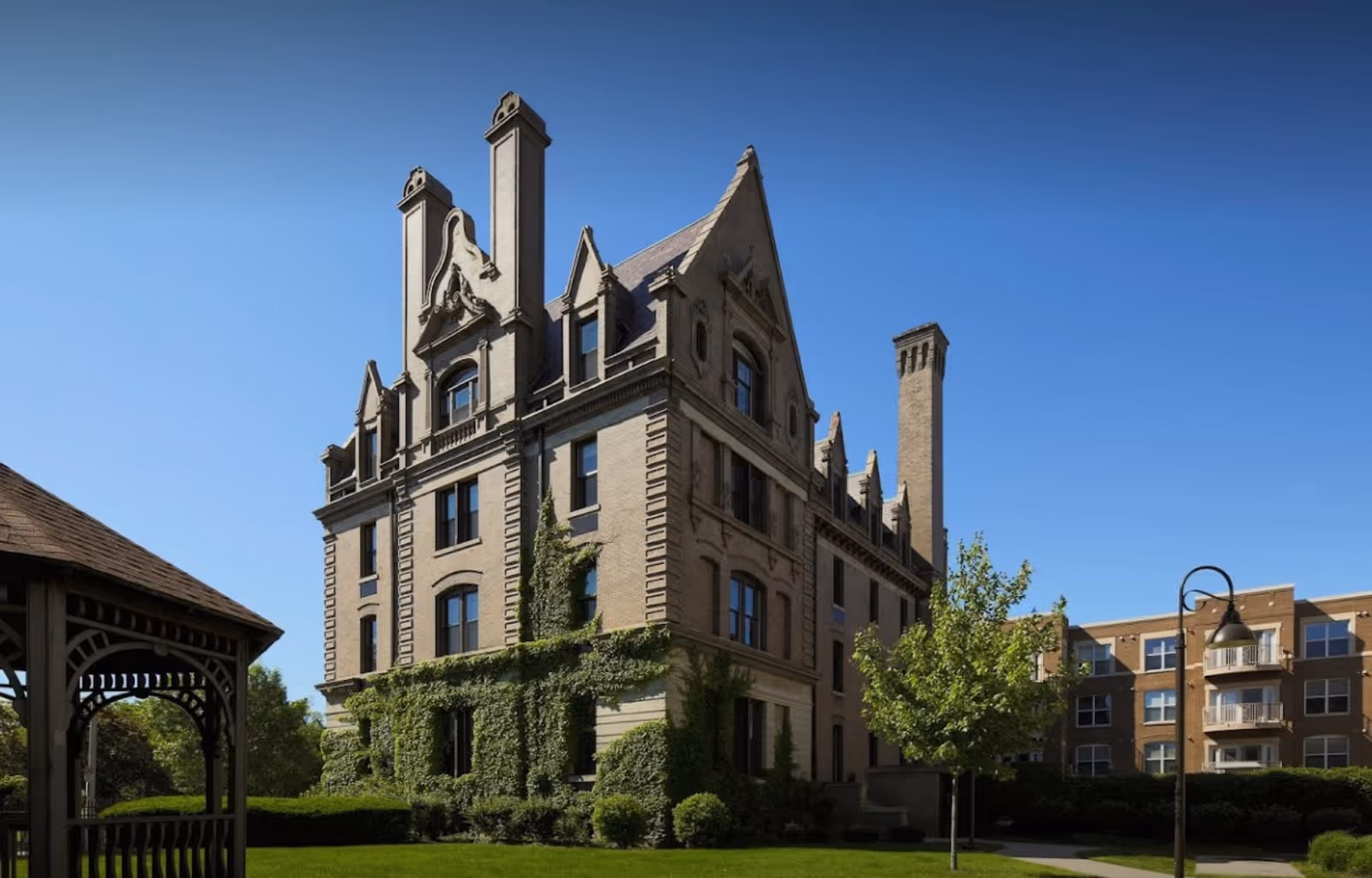 A large, historic-style brick building with ornate architectural details and tall chimneys, partially covered with green ivy, under a clear blue sky. There is a well-maintained lawn with bushes and a tree in front, and a wooden gazebo is visible on the left side. Another modern building with balconies is seen in the background on the right.