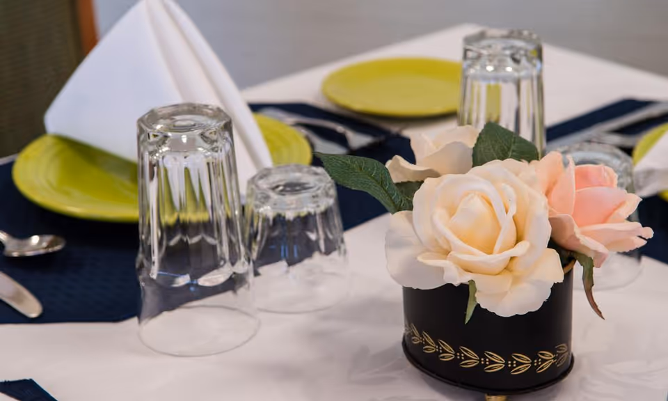 Close-up of a dining table set with upside-down clear drinking glasses, green plates, silverware, white folded napkins, and a small black vase holding light pink and cream artificial roses on a white tablecloth with dark blue placemats.