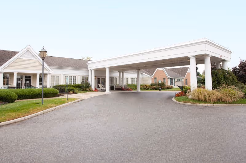 Entrance area of a senior living facility with a large covered driveway supported by white columns, surrounded by well-maintained landscaping and single-story buildings with beige siding and white trim.