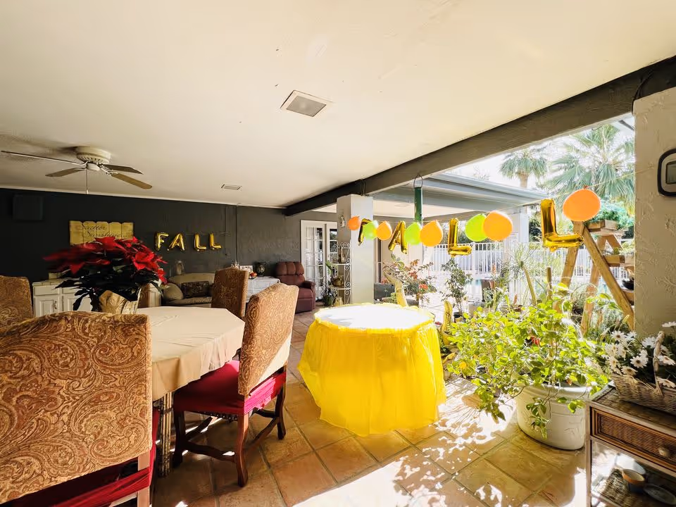 Covered outdoor patio area with a round table covered in a yellow cloth, surrounded by plants and decorated with orange and green balloons spelling out FALL. There are upholstered chairs around another table with a beige tablecloth, a ceiling fan, and a dark wall with gold balloon letters spelling FALL.