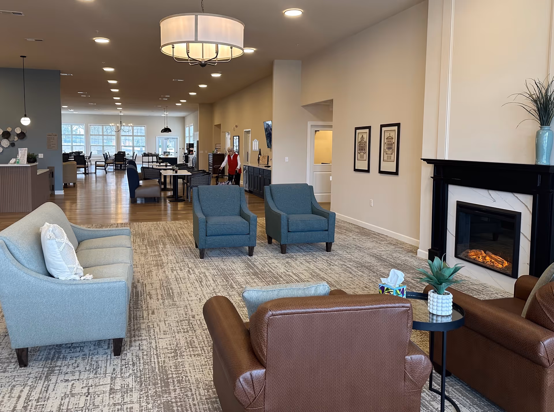 A spacious senior living facility common area with comfortable seating including a light blue sofa, two blue armchairs, and two brown armchairs around a small black table with a plant and tissue box. There is a modern electric fireplace on the right wall with a vase and plant on the mantel. The room has a patterned carpet and wooden flooring leading to a dining area with tables and chairs. A senior woman with a walker is visible in the background near the dining area. The ceiling has recessed lighting and a large round chandelier.