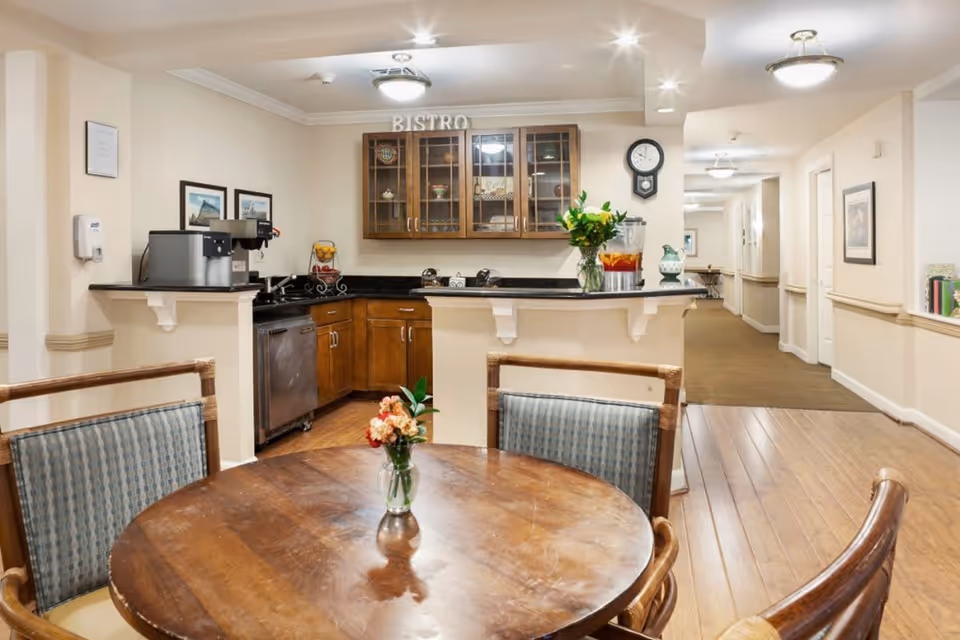 Interior view of a bistro area in a senior living facility featuring a round wooden table with four chairs, a small vase with flowers on the table, a kitchen counter with a coffee machine, a glass cabinet labeled 'BISTRO', and a hallway leading to other rooms.