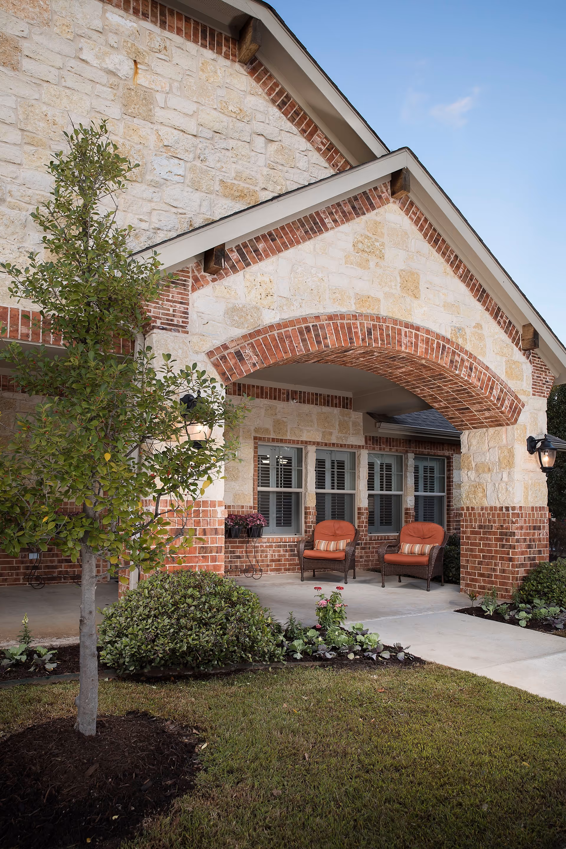 Front exterior view of a building with stone and brick facade, a covered porch with two cushioned chairs, a small tree, shrubs, and a well-maintained lawn under a clear sky.