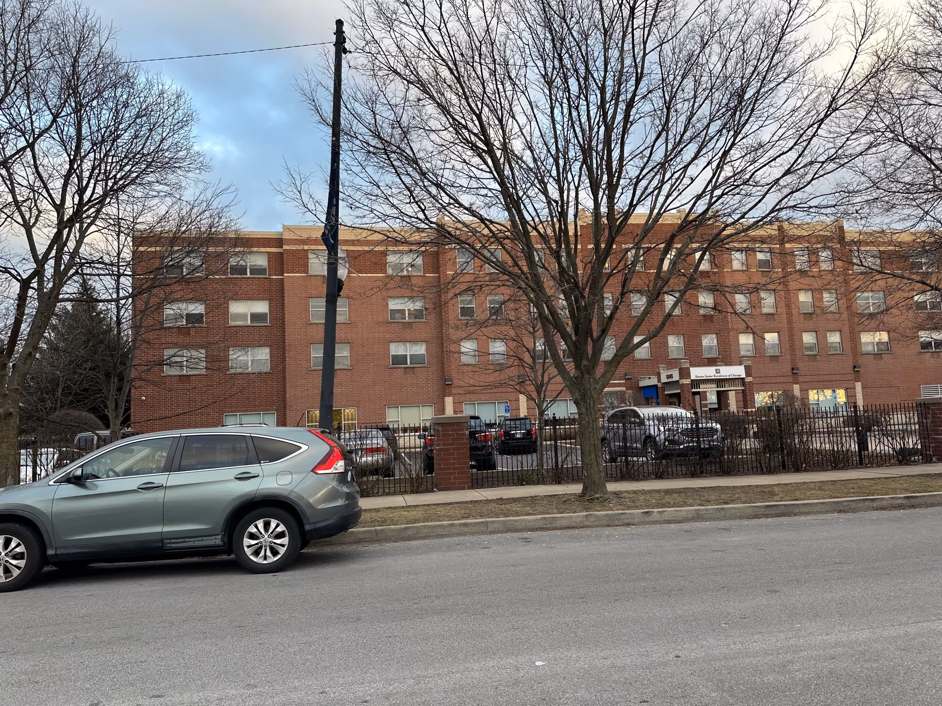Exterior view of a multi-story brick building with several windows, a parking lot with cars, leafless trees, and a sidewalk in front. The building has a sign near the entrance that reads 'Barton Senior Residences'.
