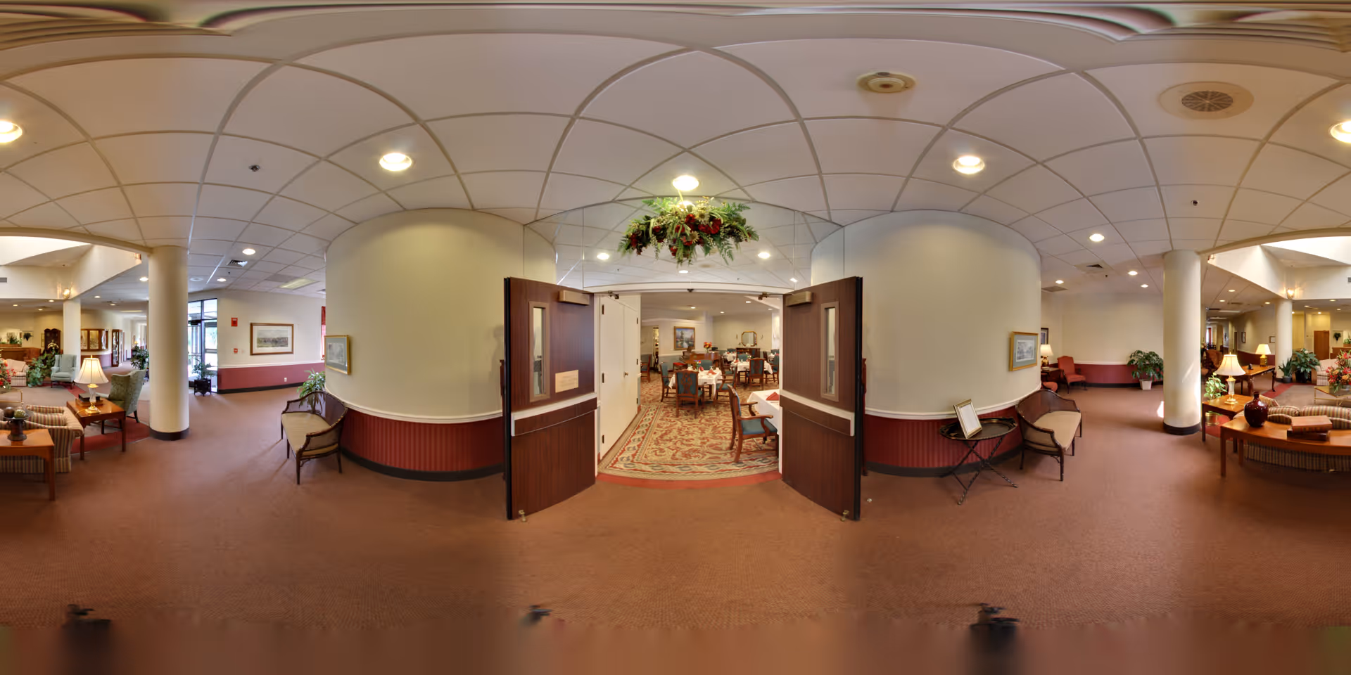 A spacious interior common area in a senior living facility with a carpeted floor, comfortable seating including armchairs and sofas, side tables with lamps, and decorative plants. Double wooden doors open into a dining room with tables and chairs arranged for meals. The ceiling has recessed lighting and a decorative floral arrangement hangs above the doorway.