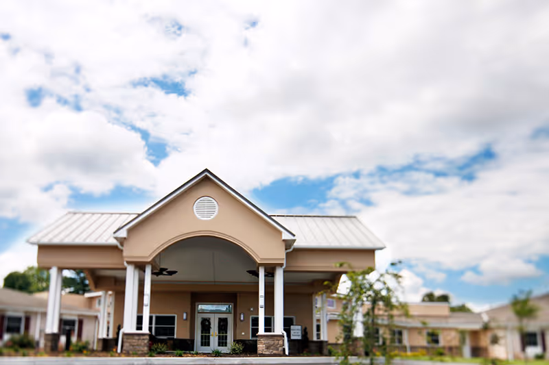 Front exterior view of a single-story retirement community building with a covered entrance supported by white columns, beige walls, and a metal roof under a partly cloudy sky.