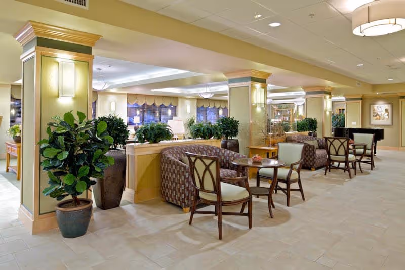 A spacious and well-lit common area in a senior living facility featuring comfortable seating arrangements with patterned armchairs and wooden chairs around small round tables. The room is decorated with large potted plants, soft yellow walls with green accents, and ceiling lights. In the background, there are windows with blinds and additional seating, creating a welcoming lounge atmosphere.