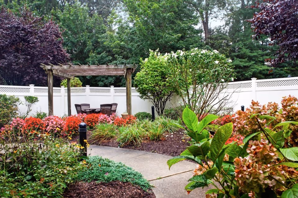 A garden area with a concrete pathway leading to a wooden pergola with a table and four chairs underneath. The garden is filled with various colorful flowers, shrubs, and green plants, surrounded by a white fence and tall trees in the background.