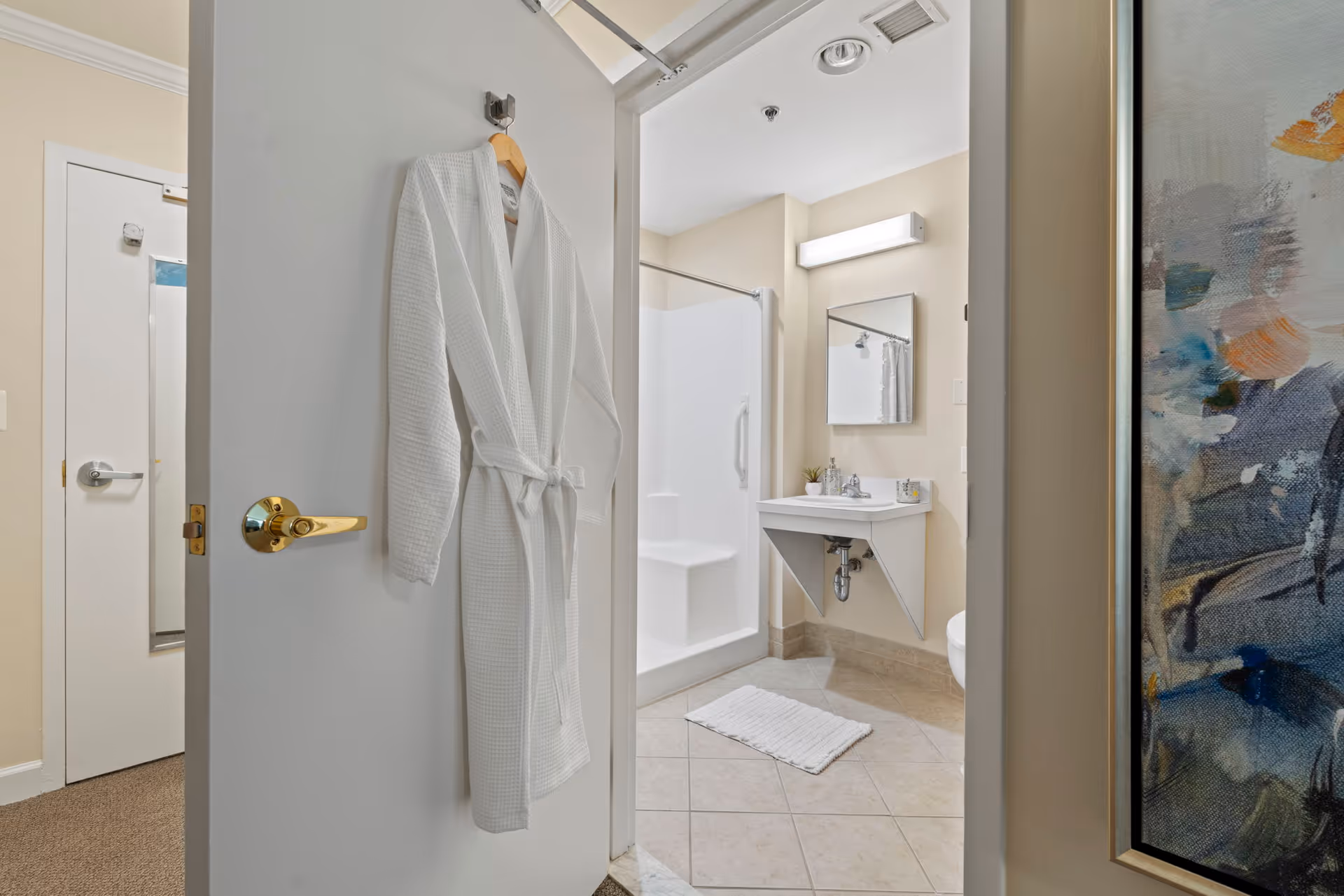 View into a bathroom from an open door with a white bathrobe hanging on the door. The bathroom features a walk-in shower with a grab bar, a wall-mounted sink with a mirror above it, a white bath mat on the tiled floor, and neutral-colored walls. Part of a colorful abstract painting is visible on the right side of the image.